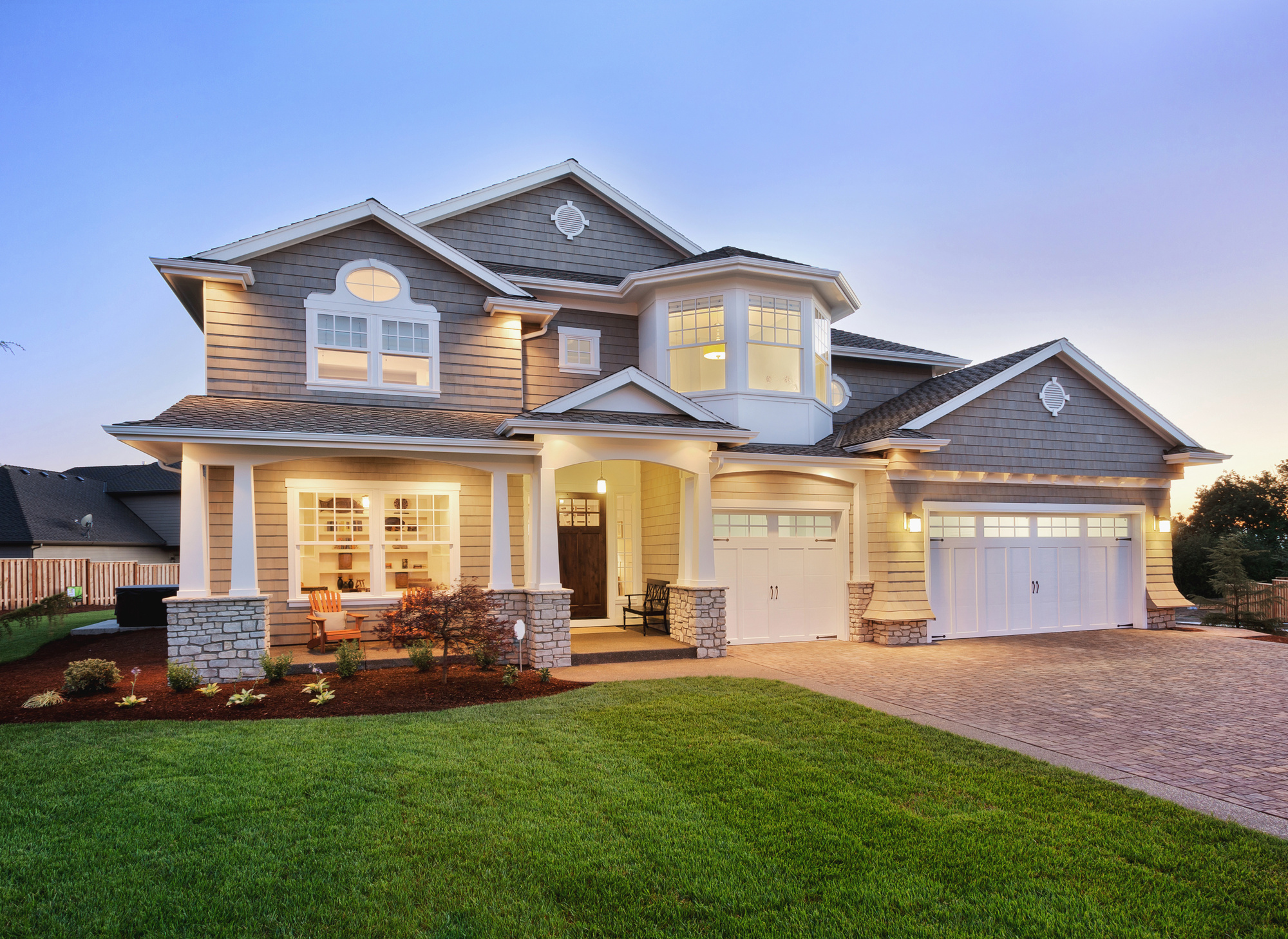 Two-story gray house with white garage doors at dusk with the lights on