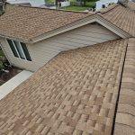 A photo of a brown and tan shingles roof from above, showing multiple sections of a suburban roof and the tan siding on the home.